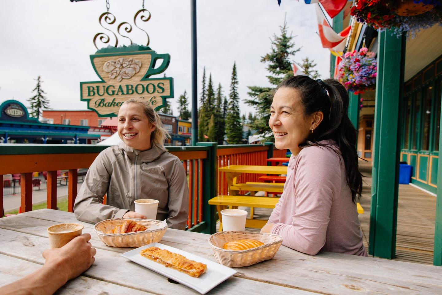 Two women sitting on an outdoor patio at Bugaboos Cafe at Silver Star Mountain having a coffee and a pastry