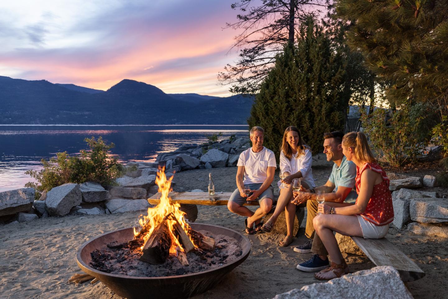 group of friends sitting around a fire pit next to the lake at sunset
