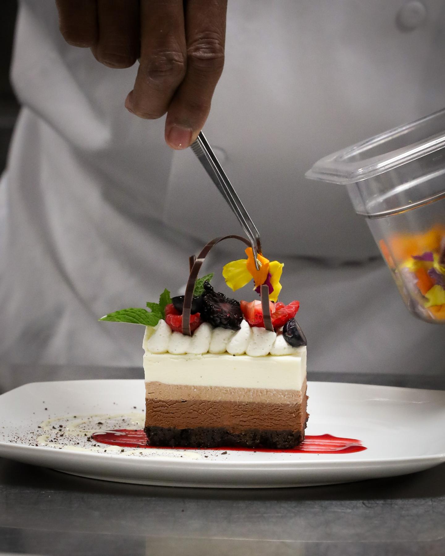 Chef garnishing a layered dessert with edible flowers and berries on a plate.