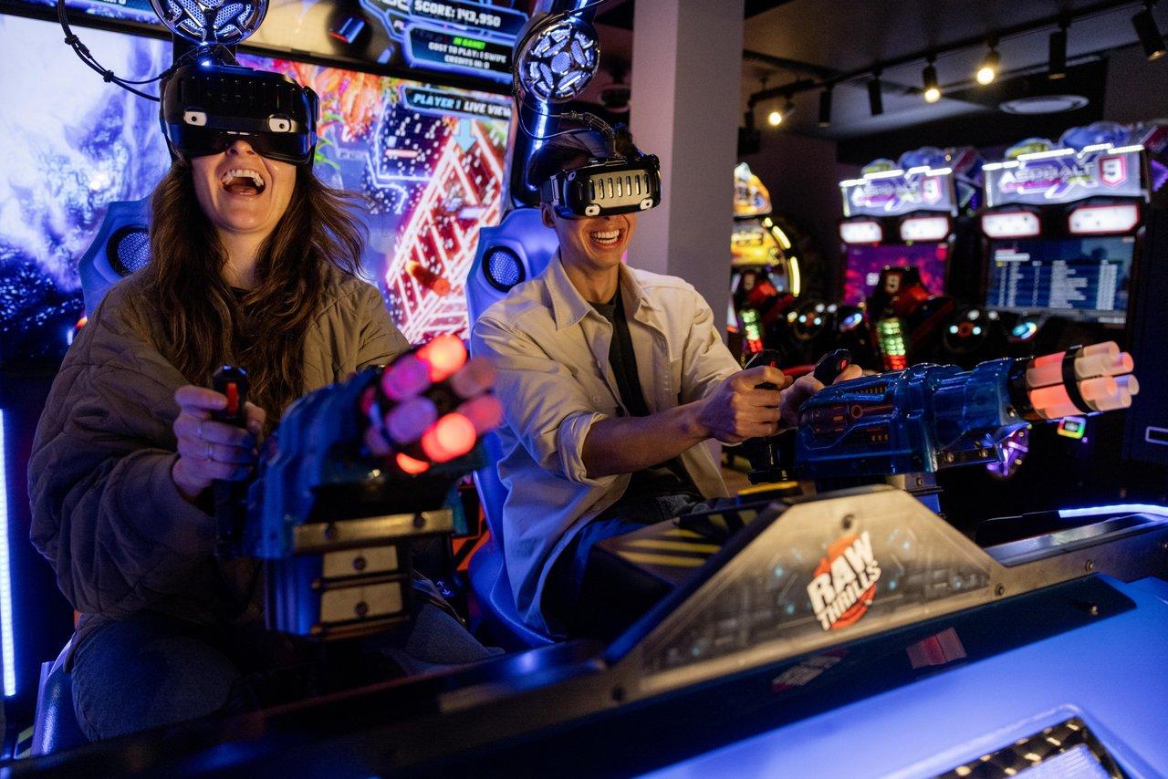 Two people playing an arcade shooting game, smiling and wearing VR headsets.