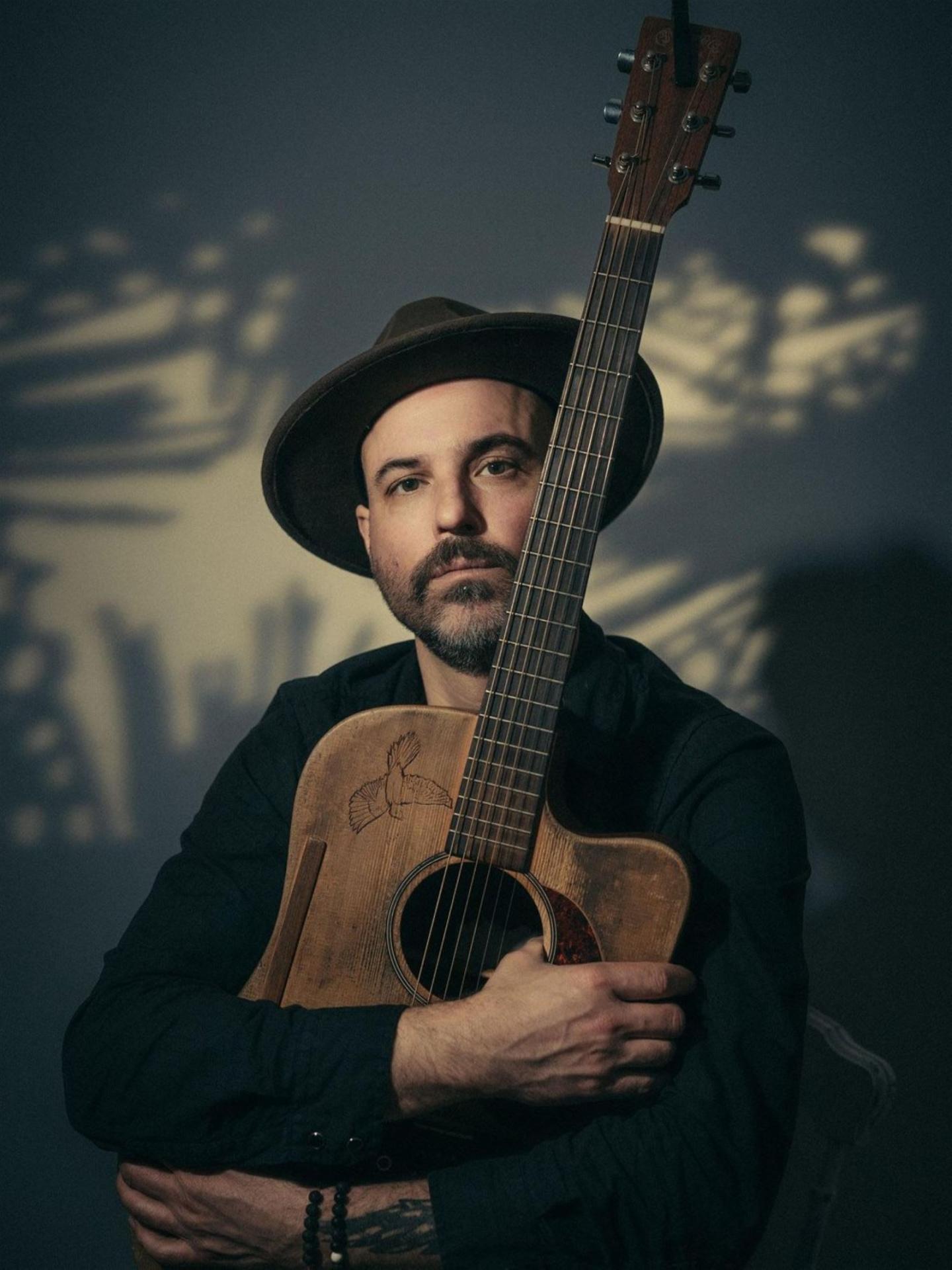 Man with hat holding an acoustic guitar, soft lighting and shadows.