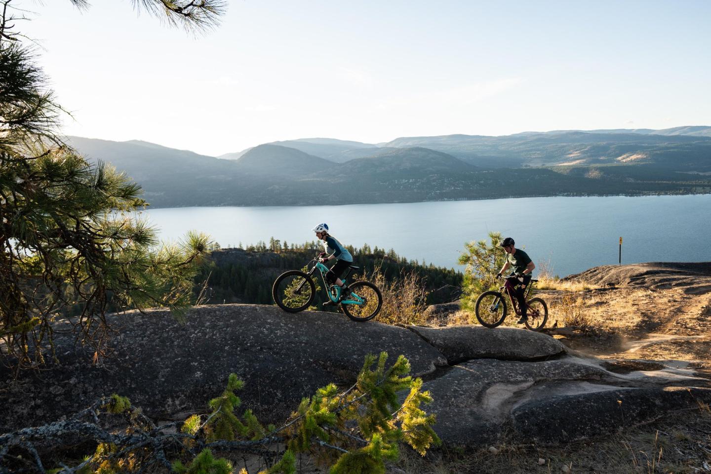 Cyclists on a rocky trail overlooking a lake and mountains.