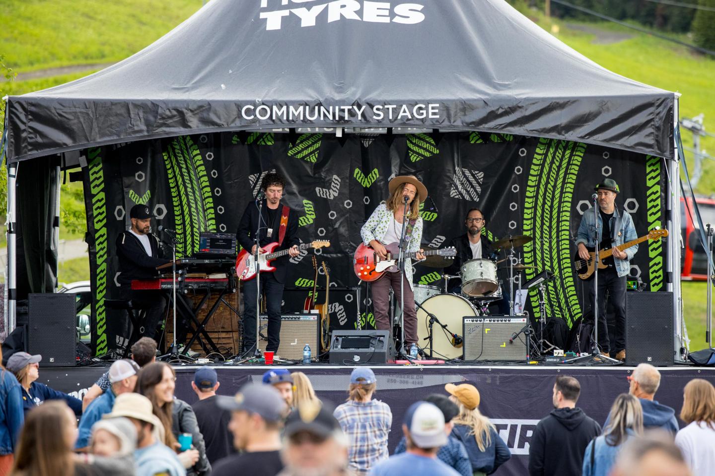 Band performing on outdoor stage at a grassy venue, with crowd watching.