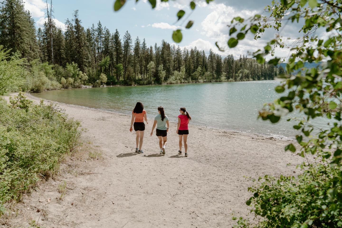 Three people walking on a lakeside beach with trees around.