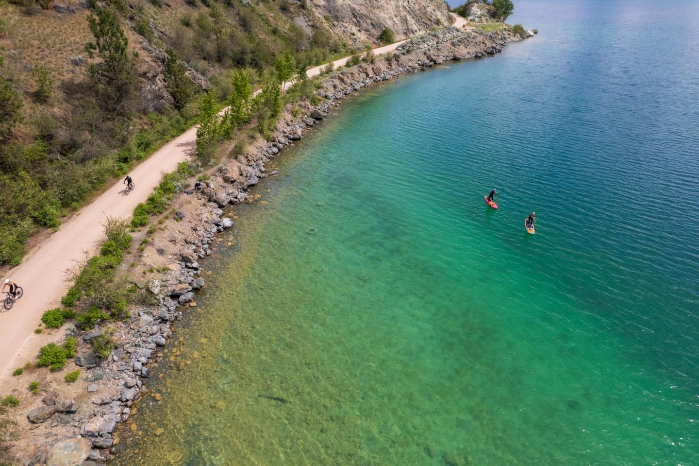 Cyclists on a path beside a turquoise lake, two kayaks on the water.