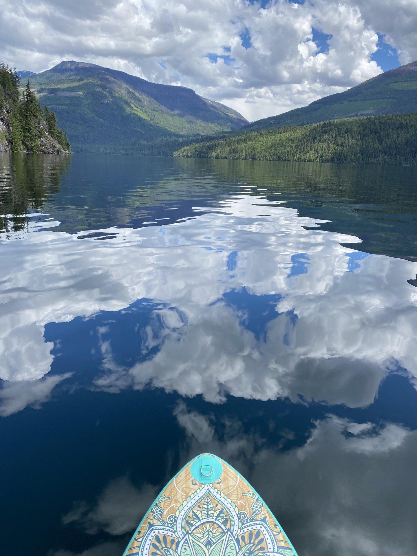 Paddleboard on calm lake reflecting mountains and clouds.