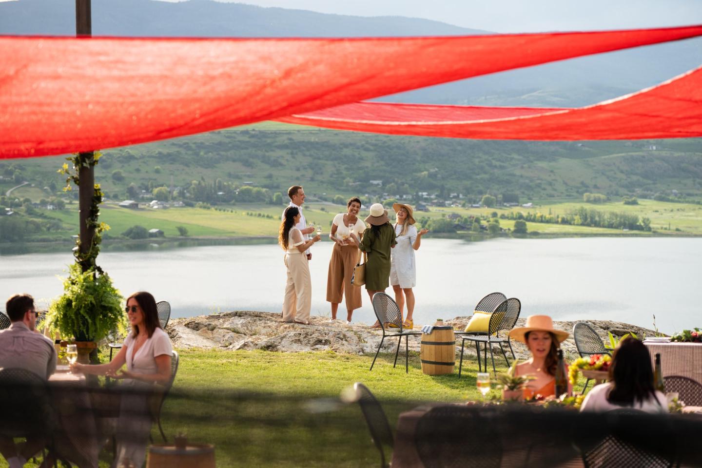 Outdoor gathering by a lake, red canopy, people seated and standing, sunny day.