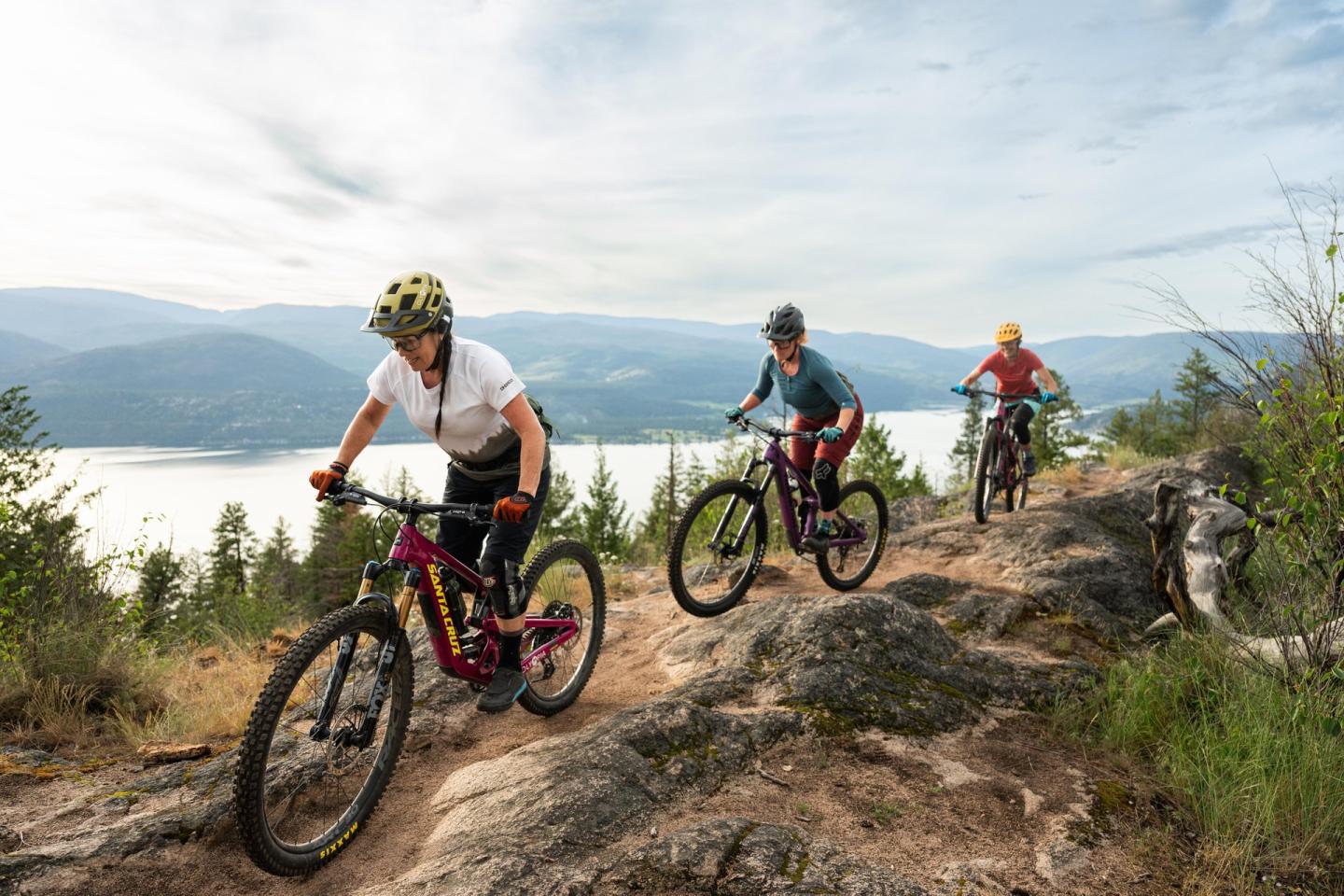 Three people mountain biking on a rocky trail overlooking a lake.