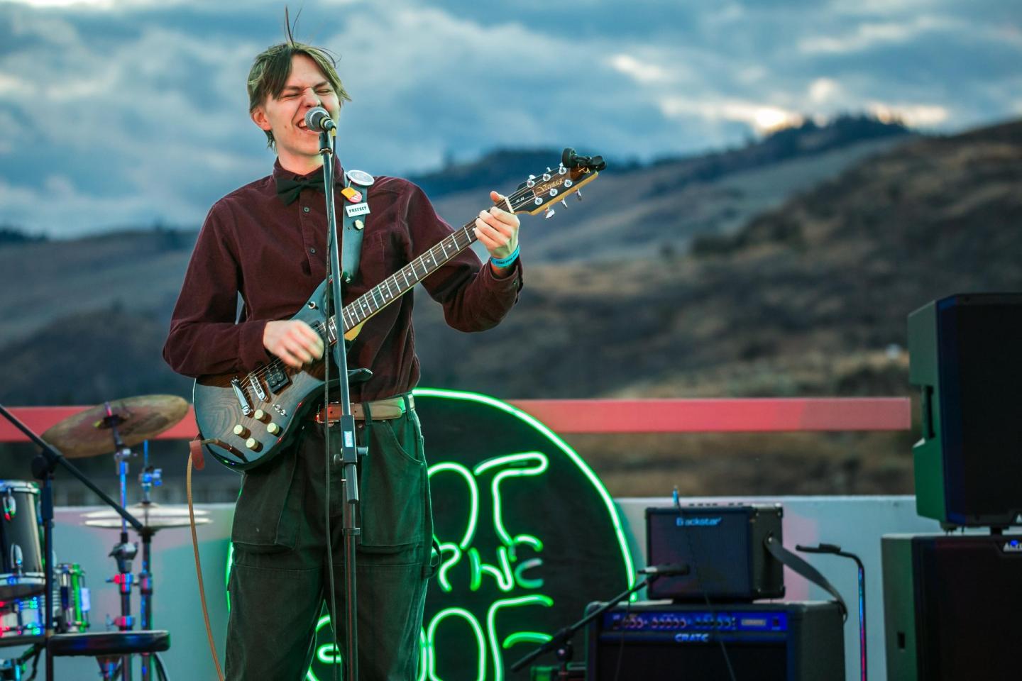 Musician playing guitar on a rooftop stage with neon sign, mountains in the background.