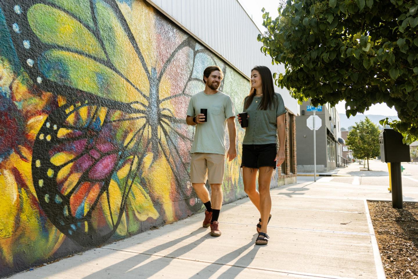 Couple walking past colorful butterfly mural on a sunny street.