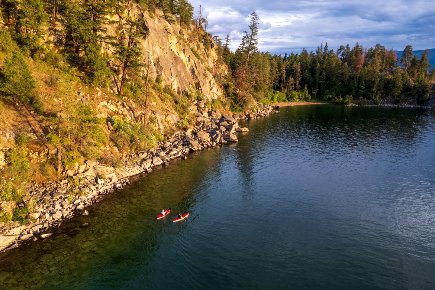 Kayakers in a serene lake beside a rocky, tree-lined cliff under a partly cloudy sky.