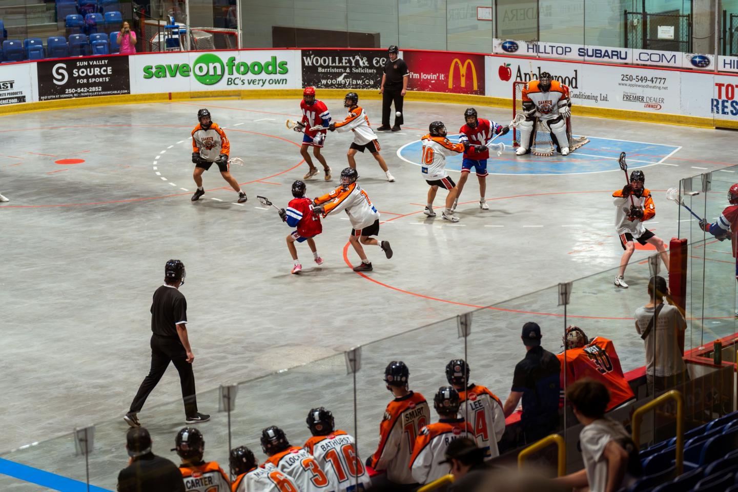 Indoor lacrosse game with players in action, orange and blue jerseys, spectators watching.