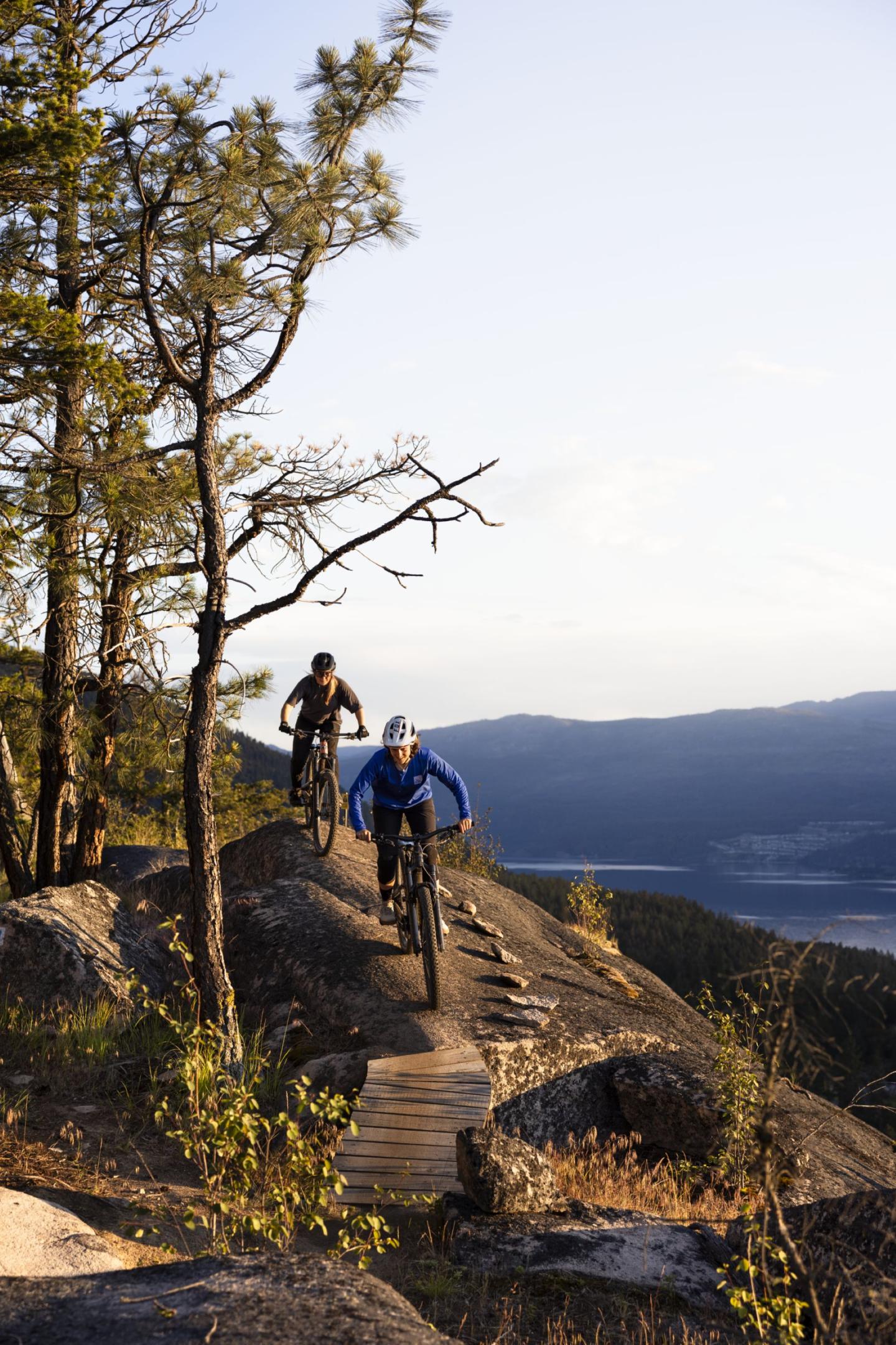 Two cyclists on a mountain trail at sunset, surrounded by trees and overlooking a valley.