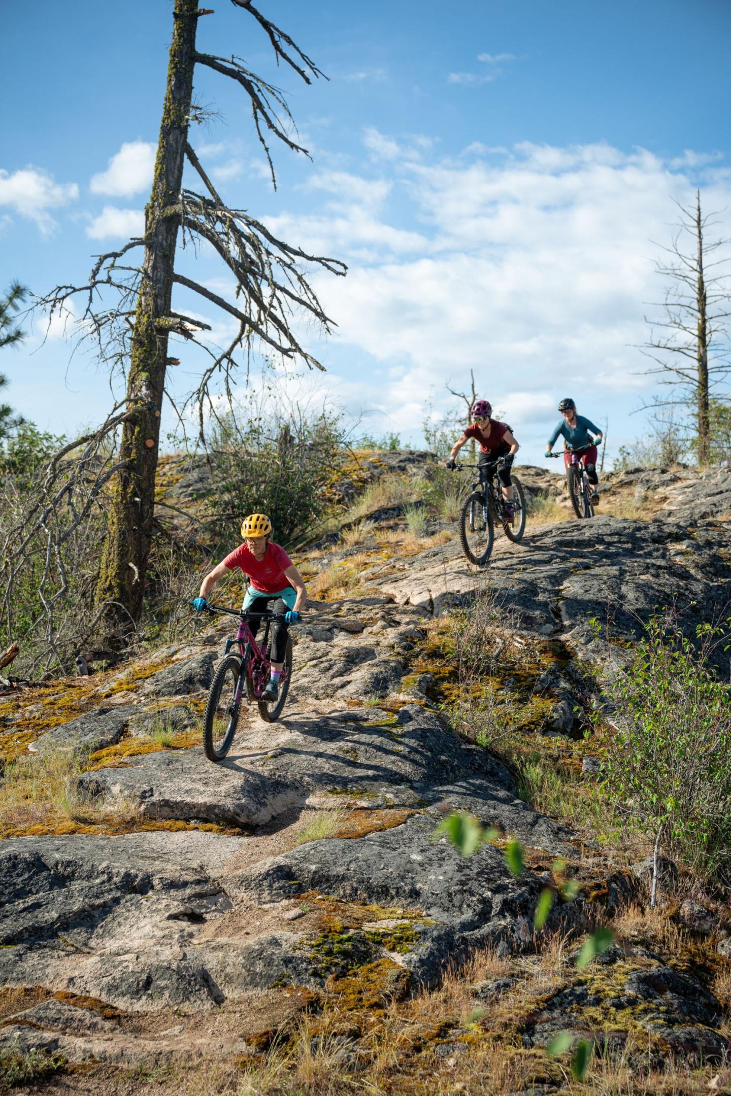 Three people mountain biking on a rocky trail under a blue sky.