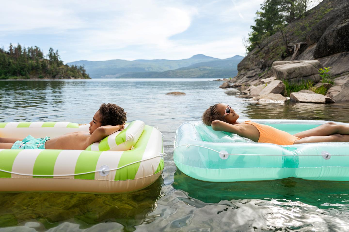 Kids lounging on pool floats in a scenic lake, surrounded by trees and mountains.