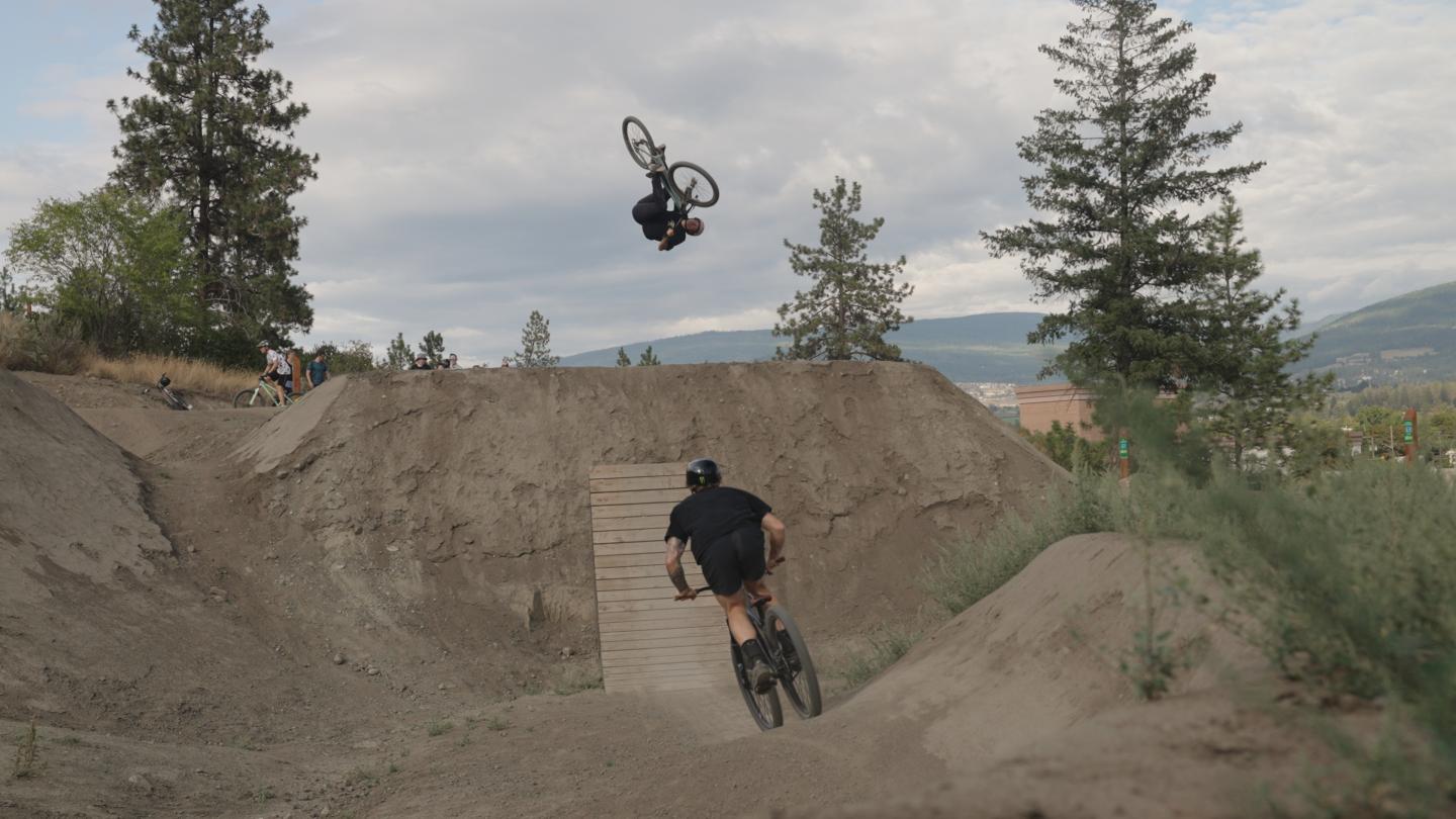 Two mountain bikers on a dirt track, one mid-air doing a flip, surrounded by trees and hills.