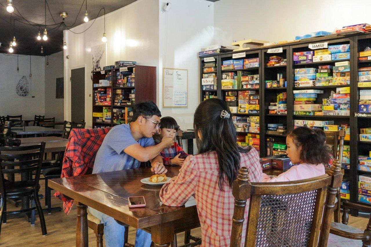 Family playing board games at a table in a cozy cafe. Shelves with games in the background.