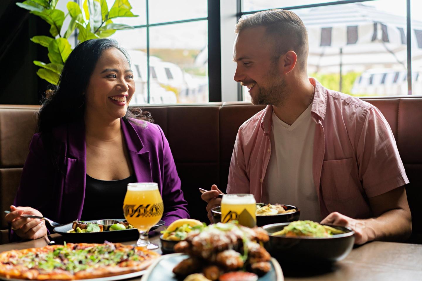 Couple smiling at each other over lunch, pizza and drinks on the table.