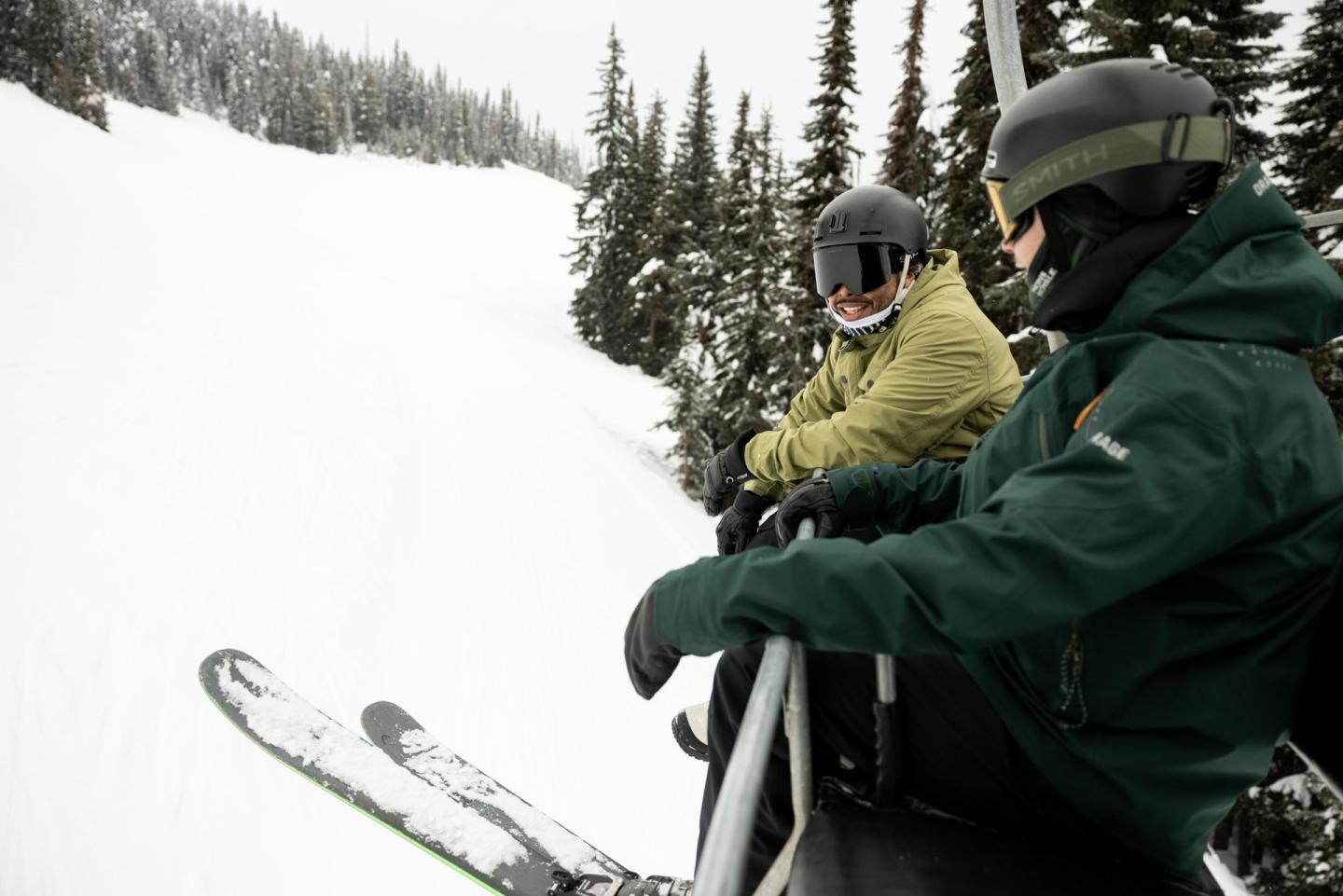 Two skiers in helmets on a ski lift, snowy trees in the background.
