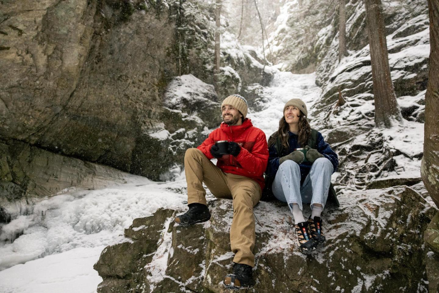 Couple in winter clothing sitting on snowy rocks in a forest, smiling.