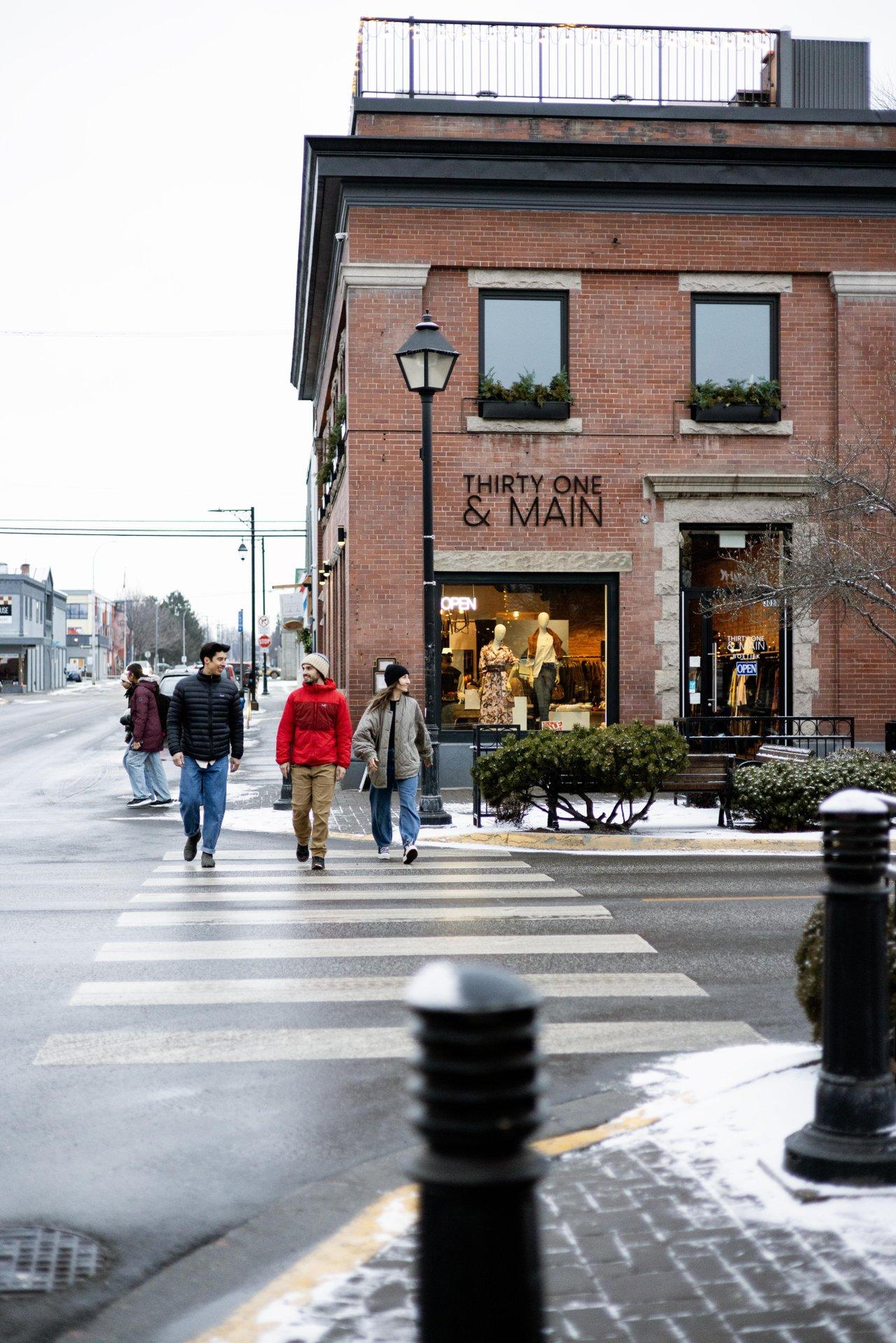 People crossing a street in front of a brick building on a cold day.