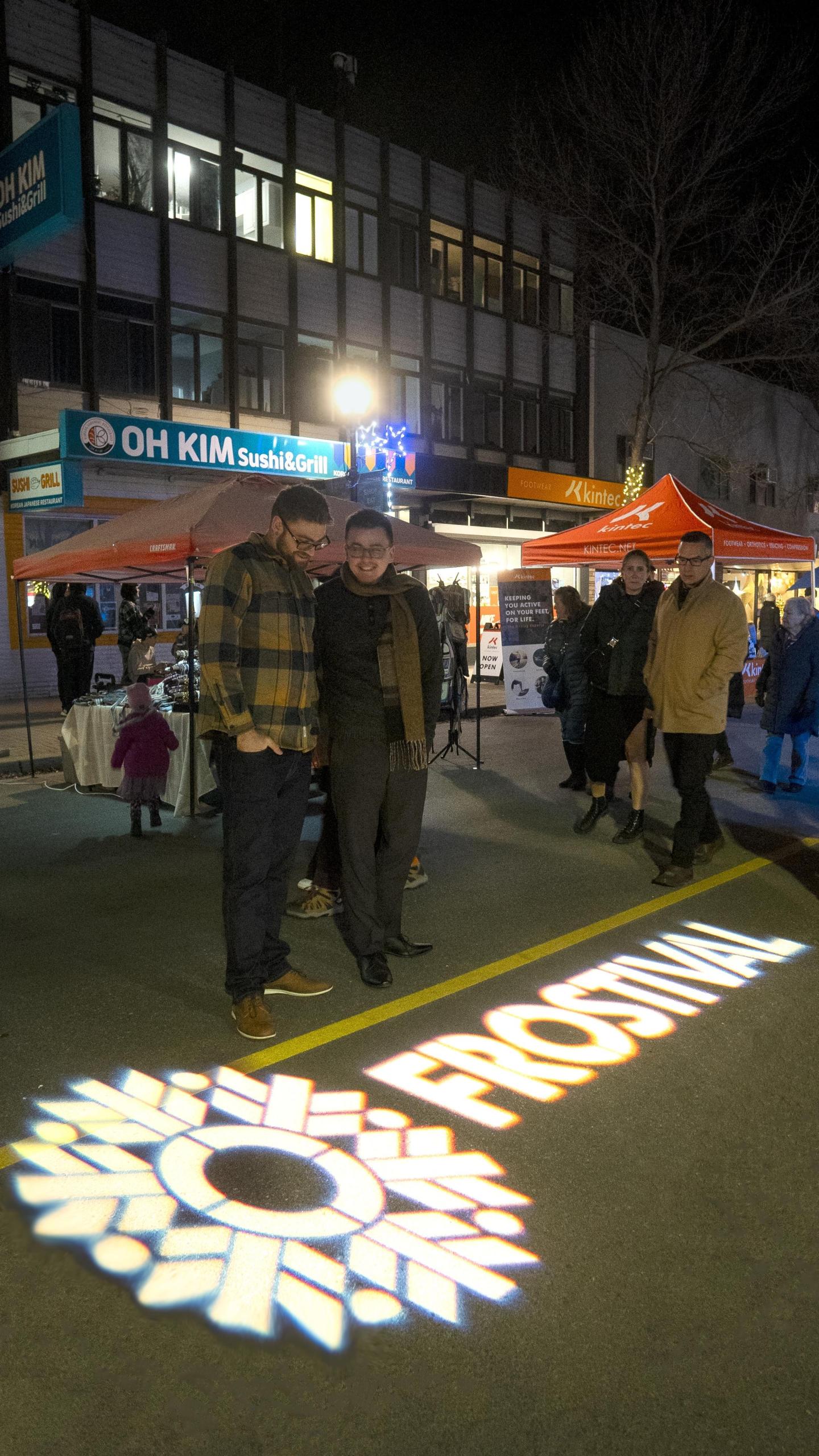 Festival-goers on street at night with colorful lights and food stalls.