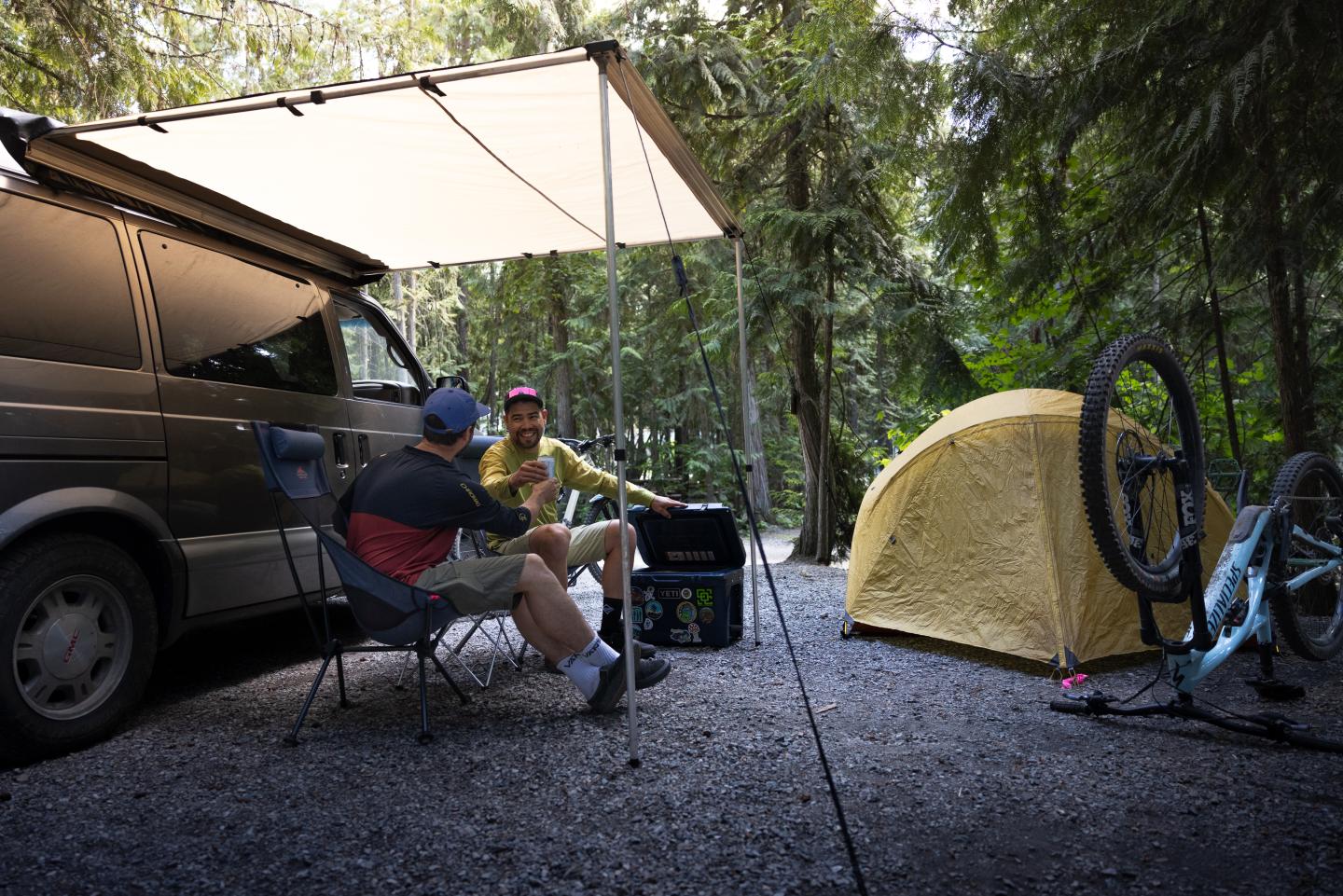 Campervan setup with awning, two people chatting, yellow tent, and bicycle in forest setting.