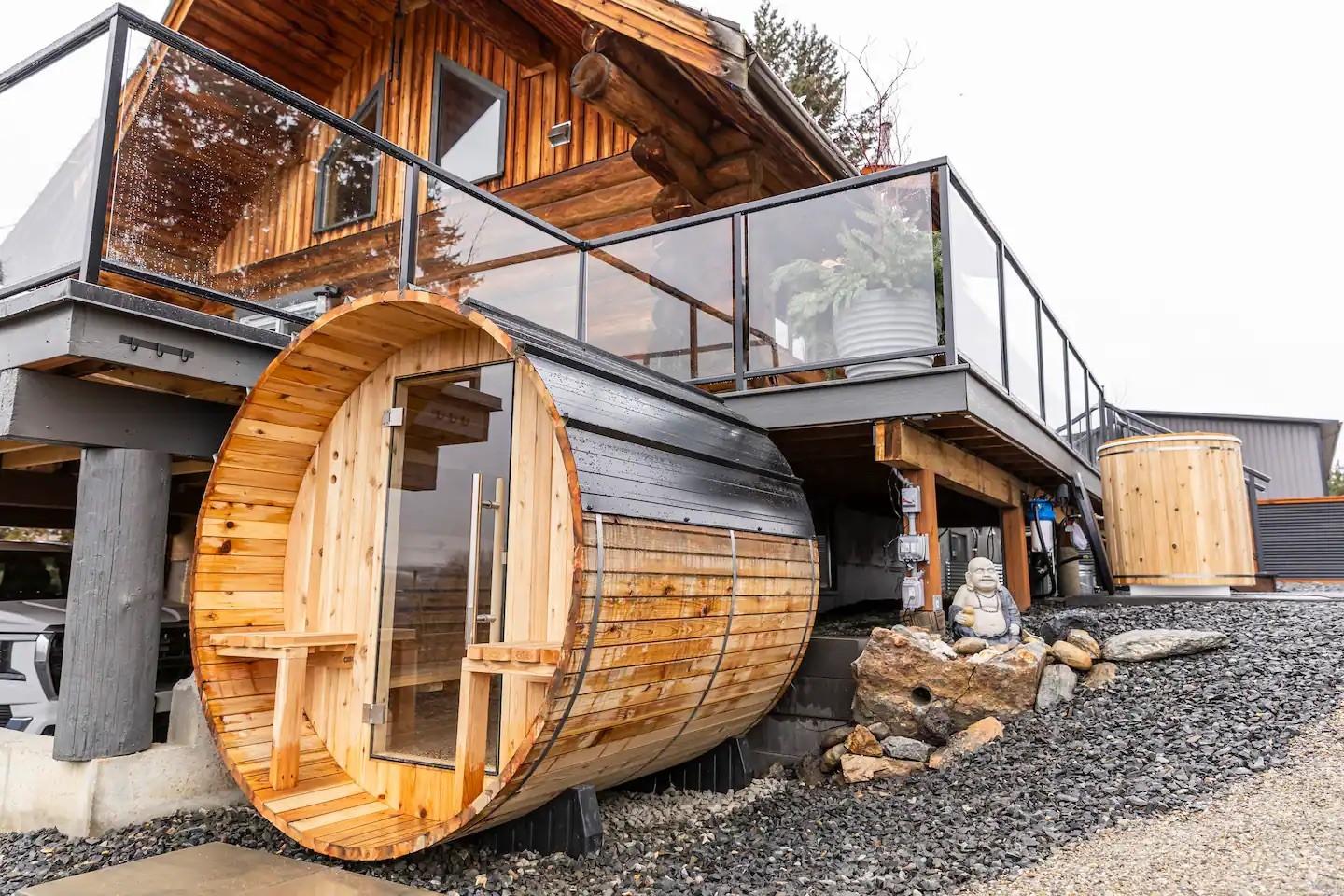 Wooden barrel sauna by modern house with deck and glass railing.