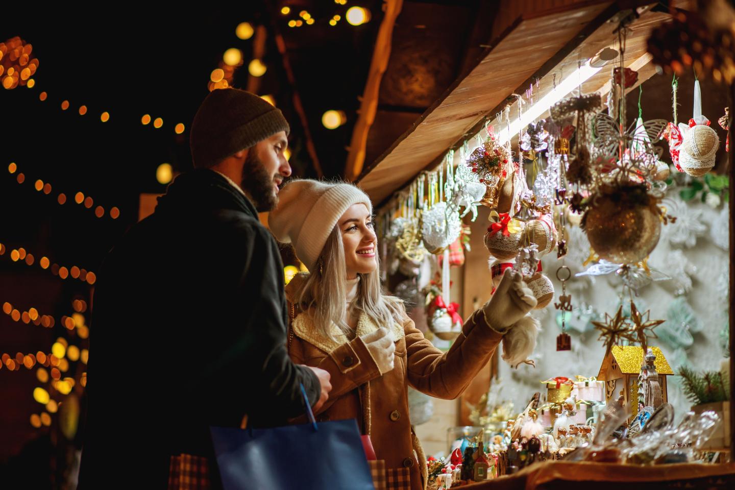A couple wearing winter coats and beanies browse festive ornaments at an outdoor Christmas market, surrounded by warm string lights and holiday decorations