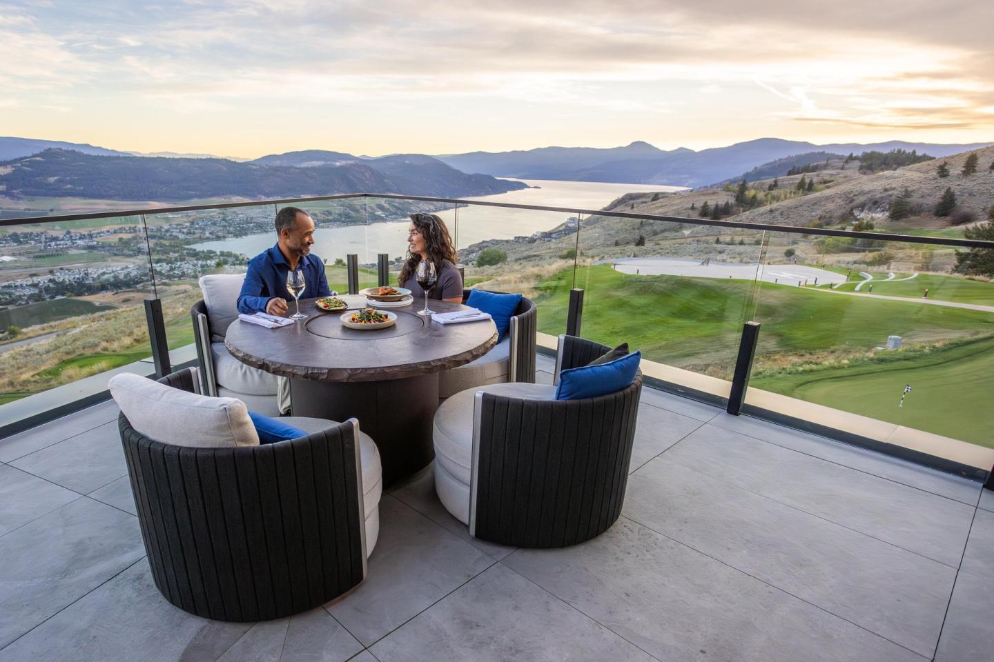 Couple dining on a patio overlooking a scenic lake and mountain landscape at sunset.