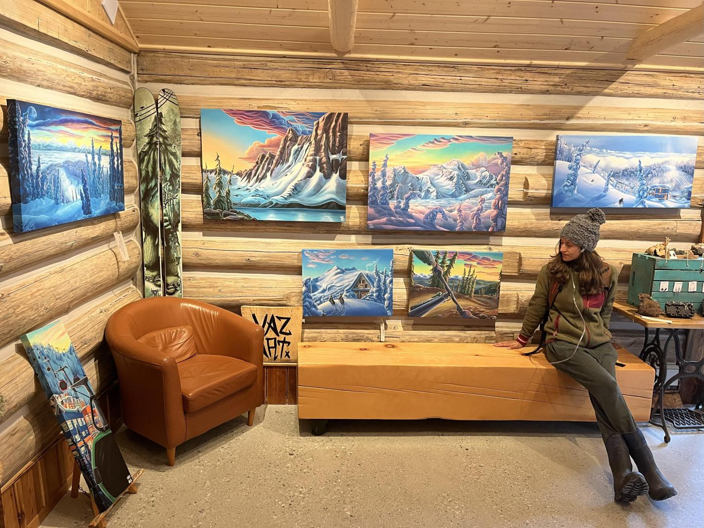 Cozy log cabin art gallery with winter landscape paintings and a seated person.