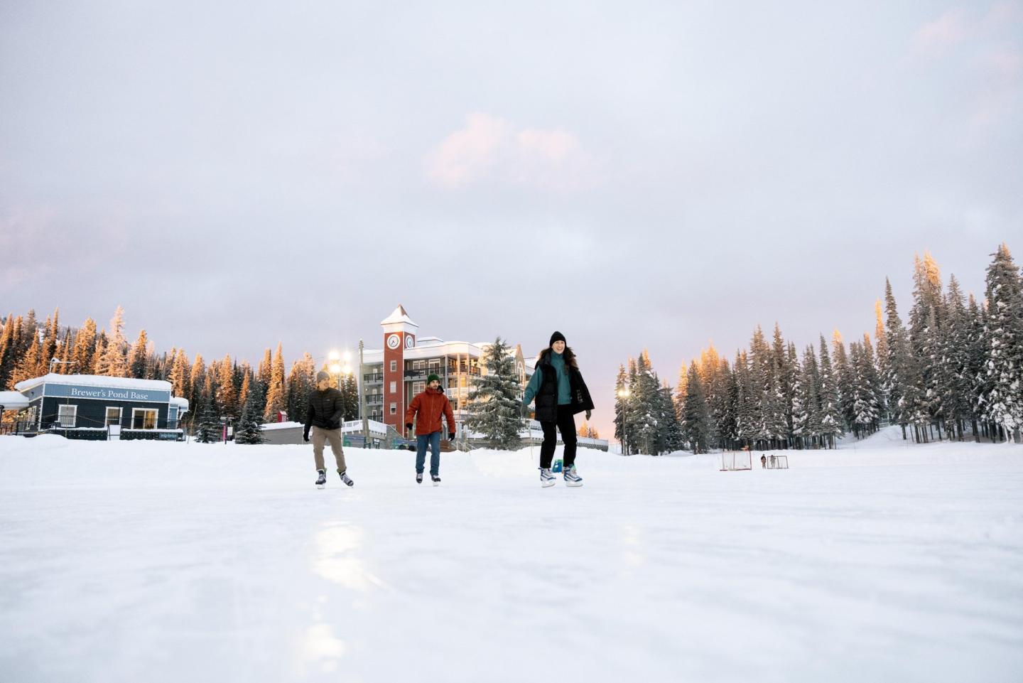 Three people ice skating on a snowy rink, with trees and buildings in the background.