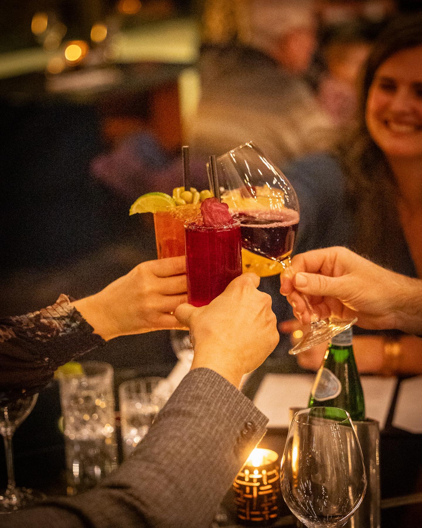 Hands toasting with colorful drinks in a dimly lit restaurant.