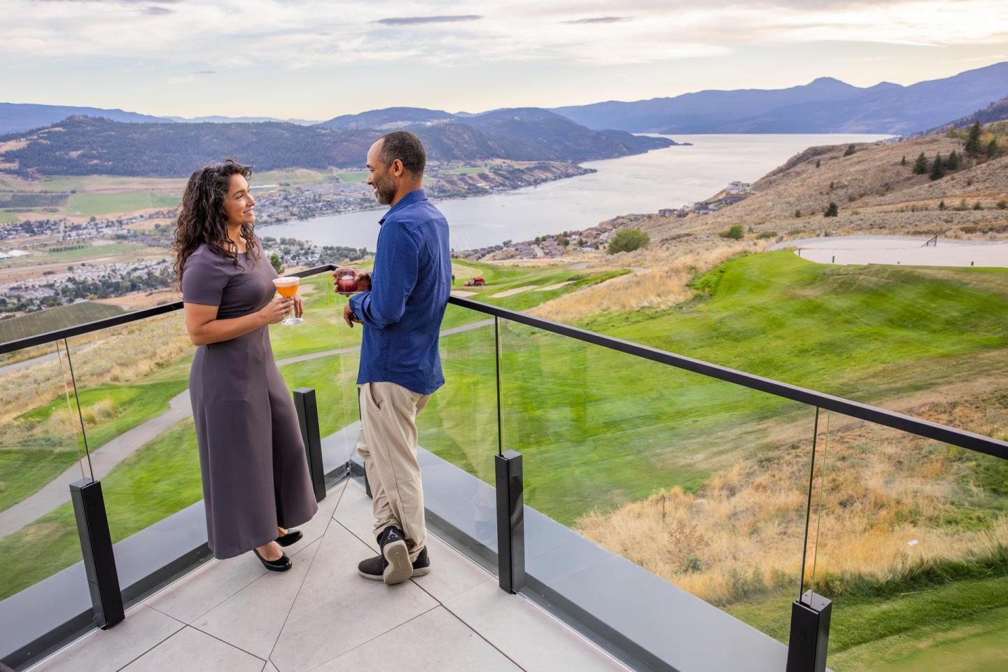 Couple enjoying drinks on a balcony, overlooking a scenic lakeside valley.