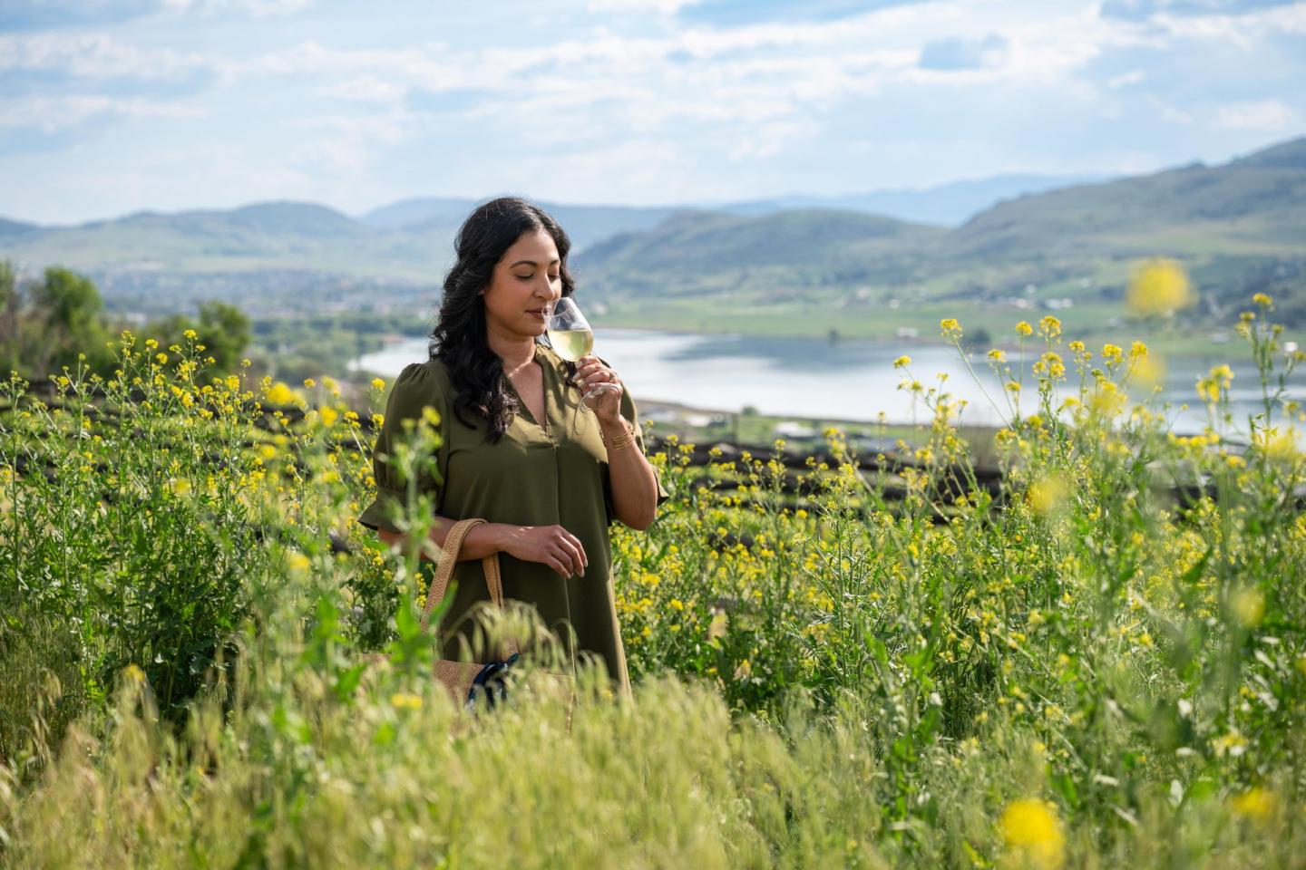 Woman in a green dress holding a drink, standing in a flowery field with hills in the background.
