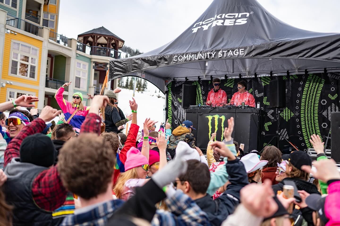 Crowd dancing at an outdoor event with DJs on stage under a canopy.