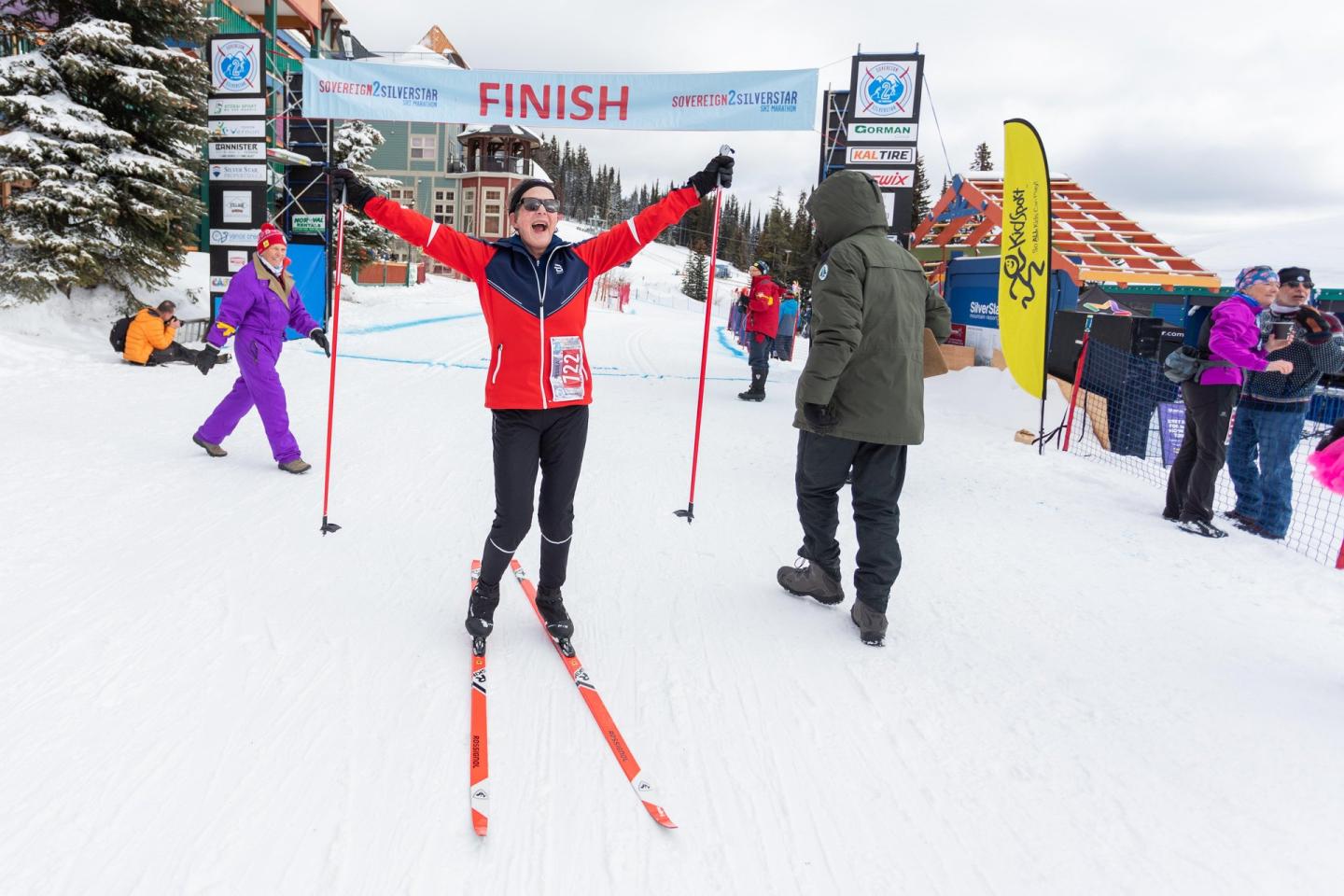Cross-country skier jubilantly finishes a race under a snowy finish line banner.