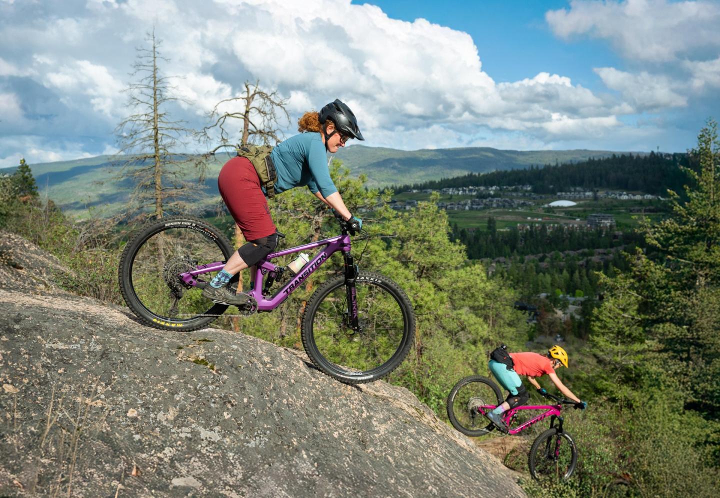Two cyclists in helmets ride down a rocky hill, forest in the background.