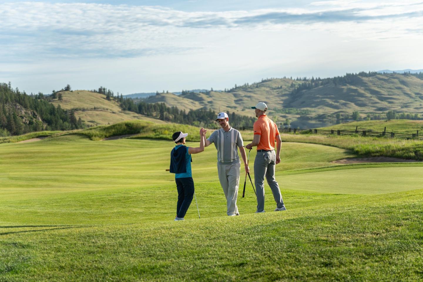 Three golfers with clubs walk and chat on a lush, sunny golf course.