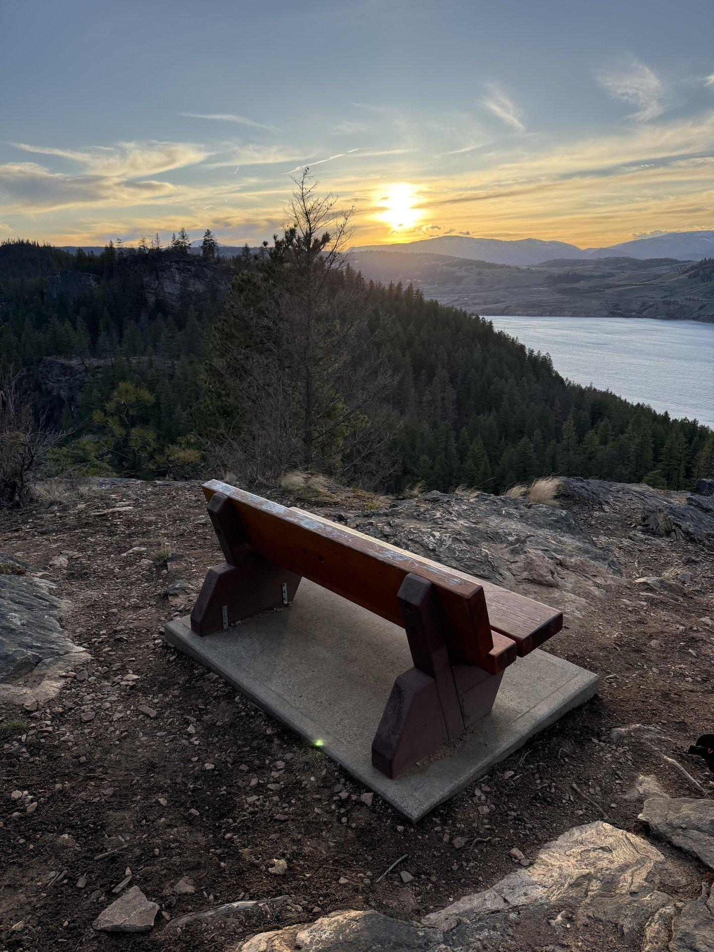 Bench on rocky hilltop overlooking lake and sunset.