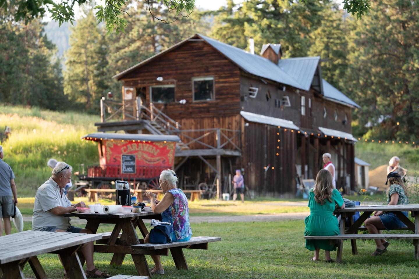People sitting at picnic tables near a rustic wooden building in a sunny, grassy area.