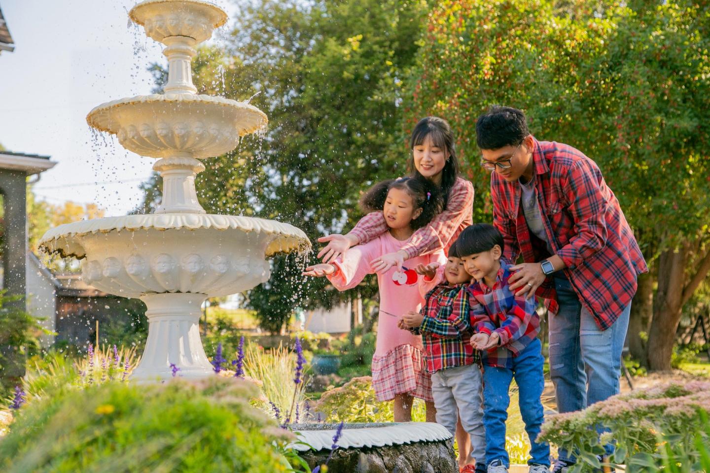 Family by a tiered fountain in a sunny garden, smiling and playing with water.