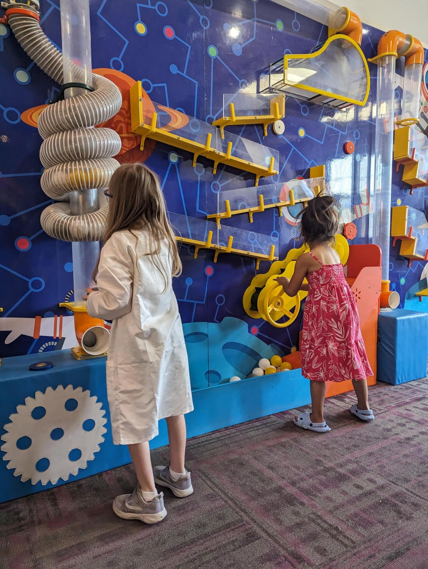 Children playing with an interactive wall exhibit featuring tubes and gears.