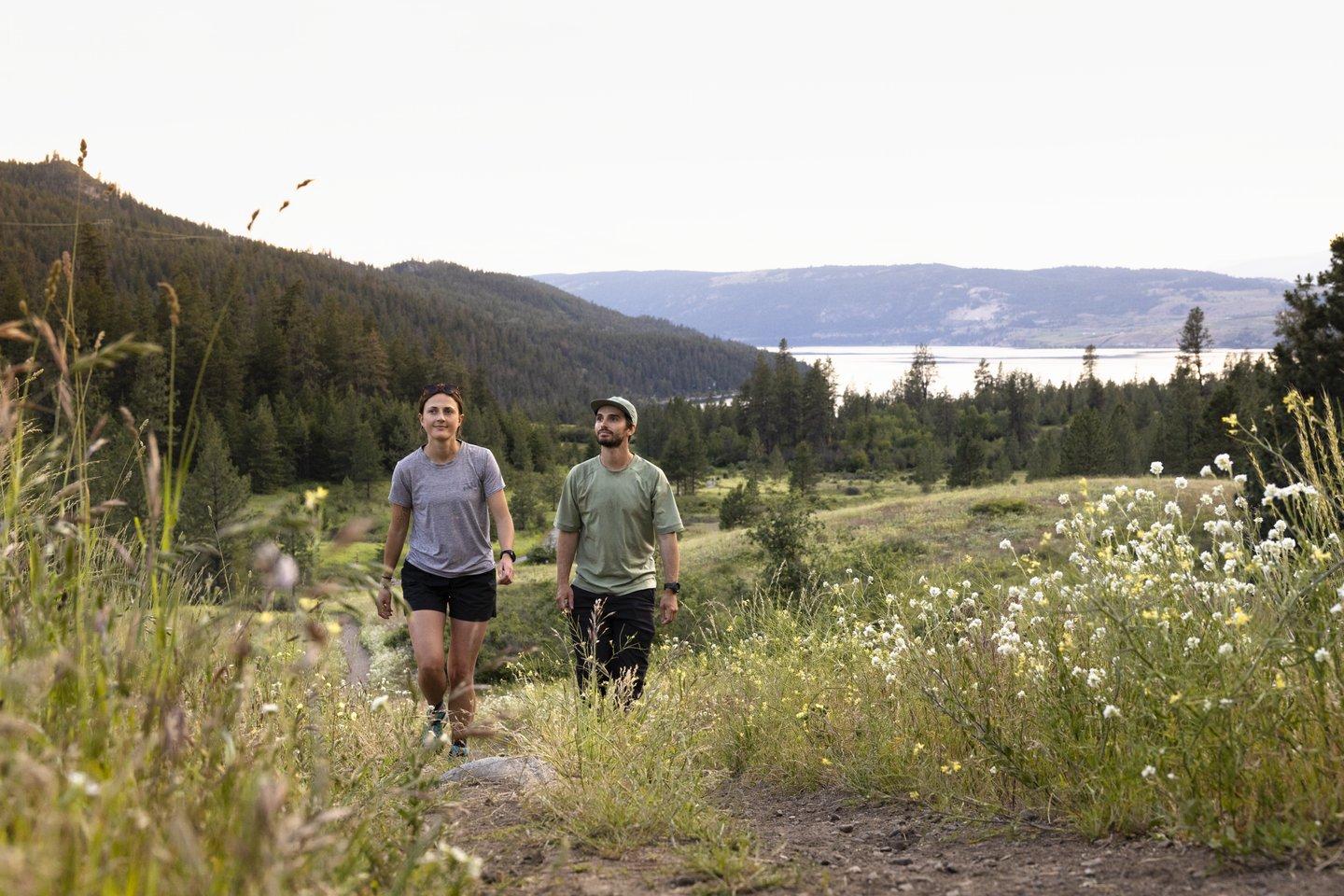 Two people hike on a grassy trail with mountains and a lake in the background.