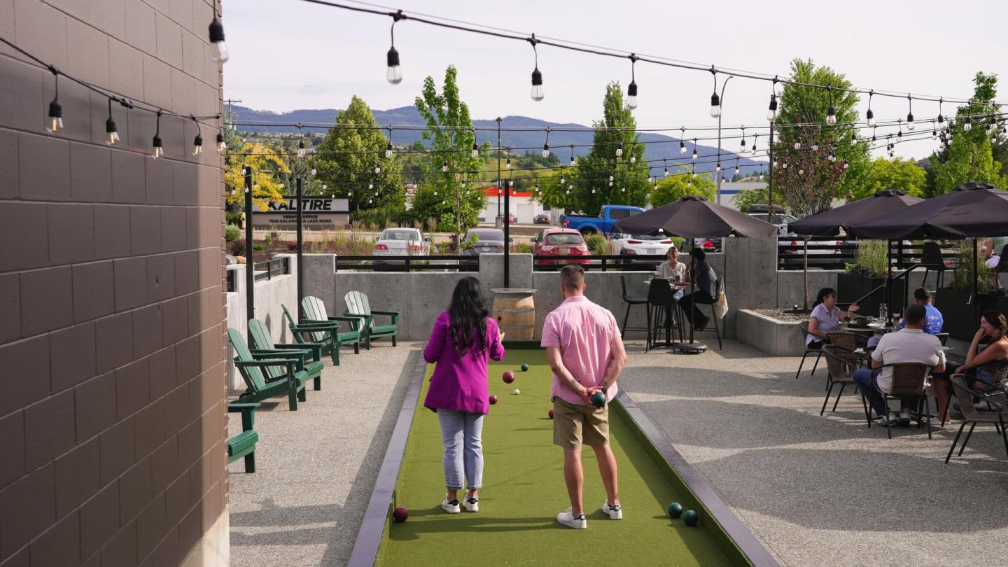 Couple playing bocce under string lights, with seated people nearby and mountains in the background.