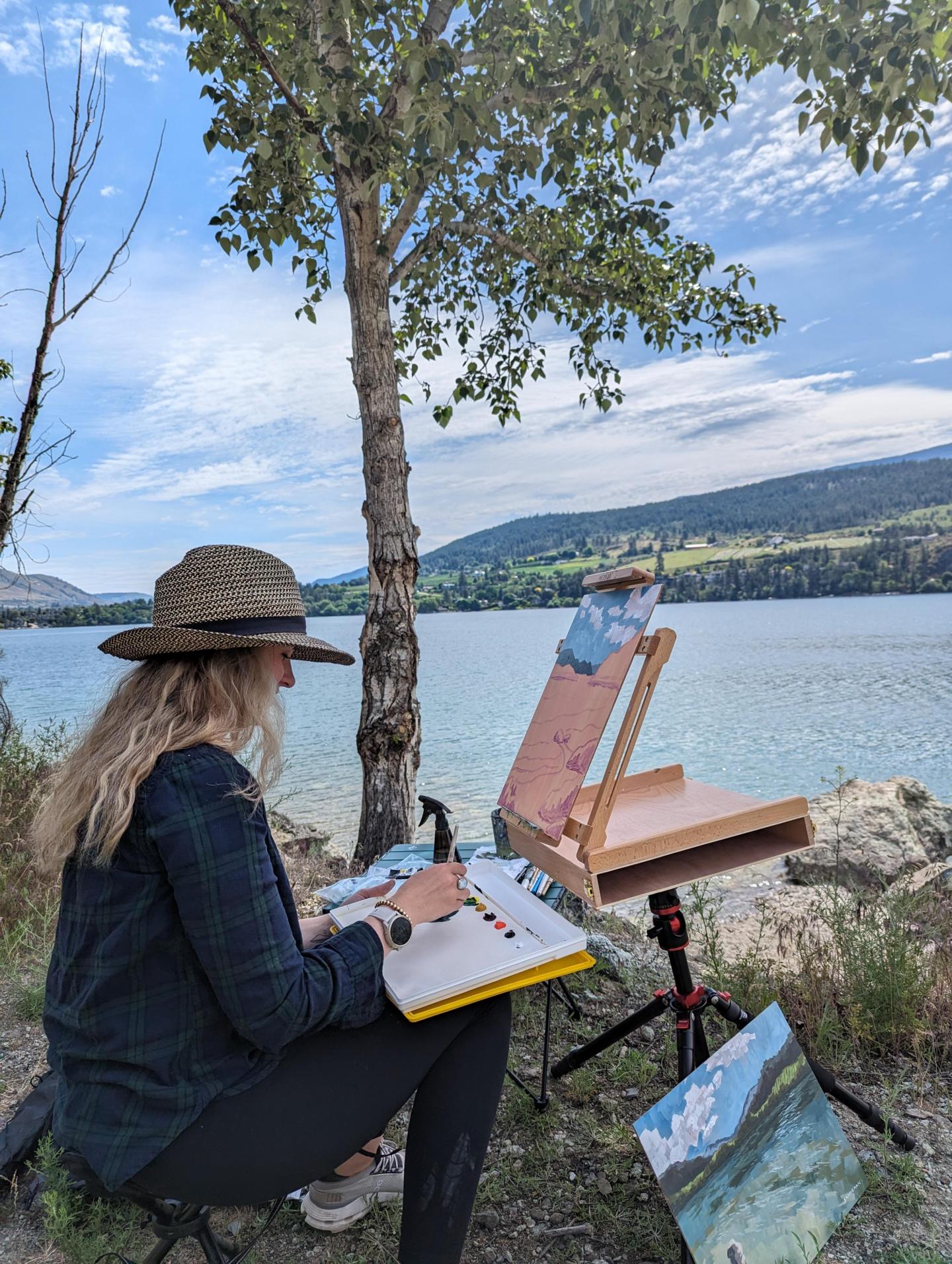 An artist sits under the shade of a tree and creates a landscape painting of the lake.