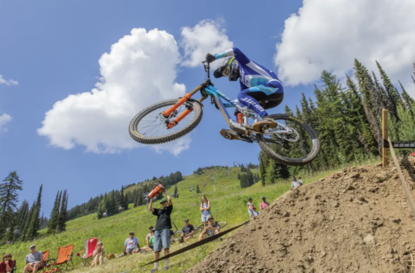 Mountain biker mid-air jump on dirt track, blue sky, and green hillside.