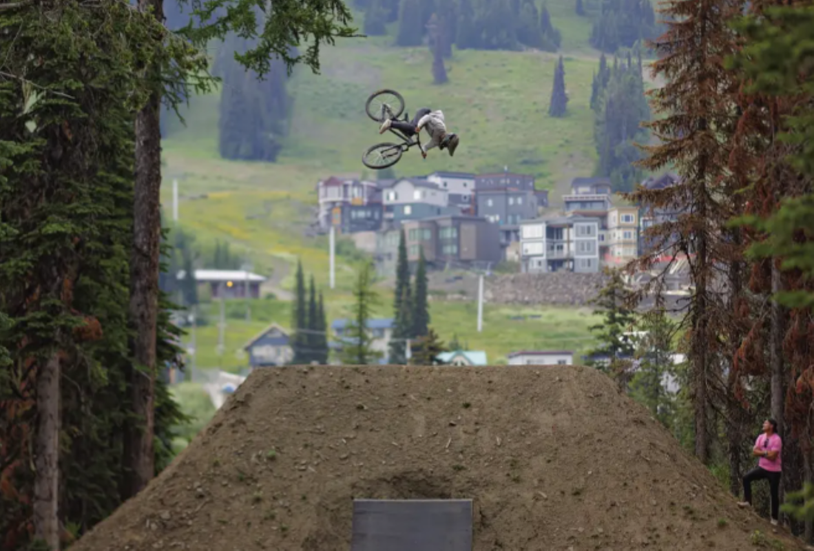 Biker performing a backflip mid-air on a dirt track in a forested area with buildings in the background.
