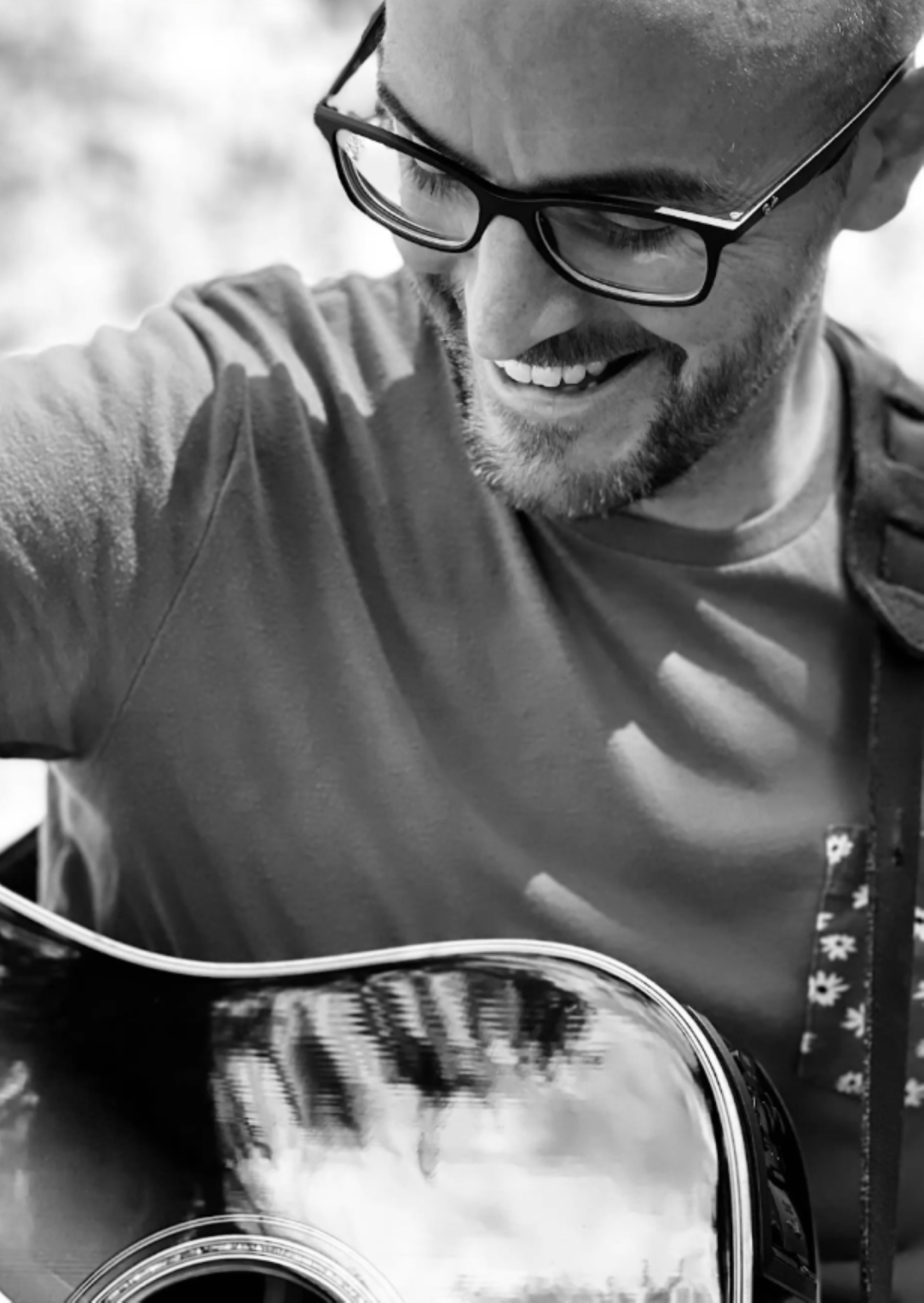 Man smiling while playing an acoustic guitar, wearing glasses.