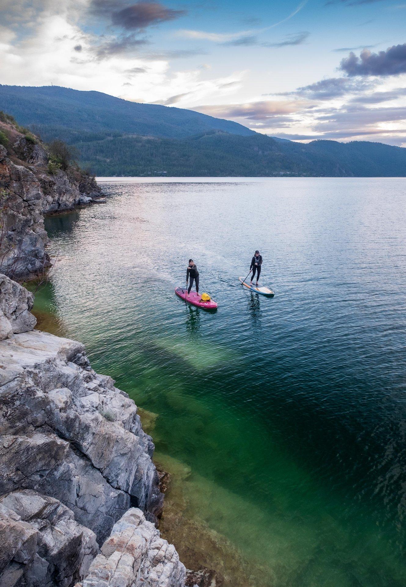 Two people paddleboarding on a calm lake near a rocky shore.